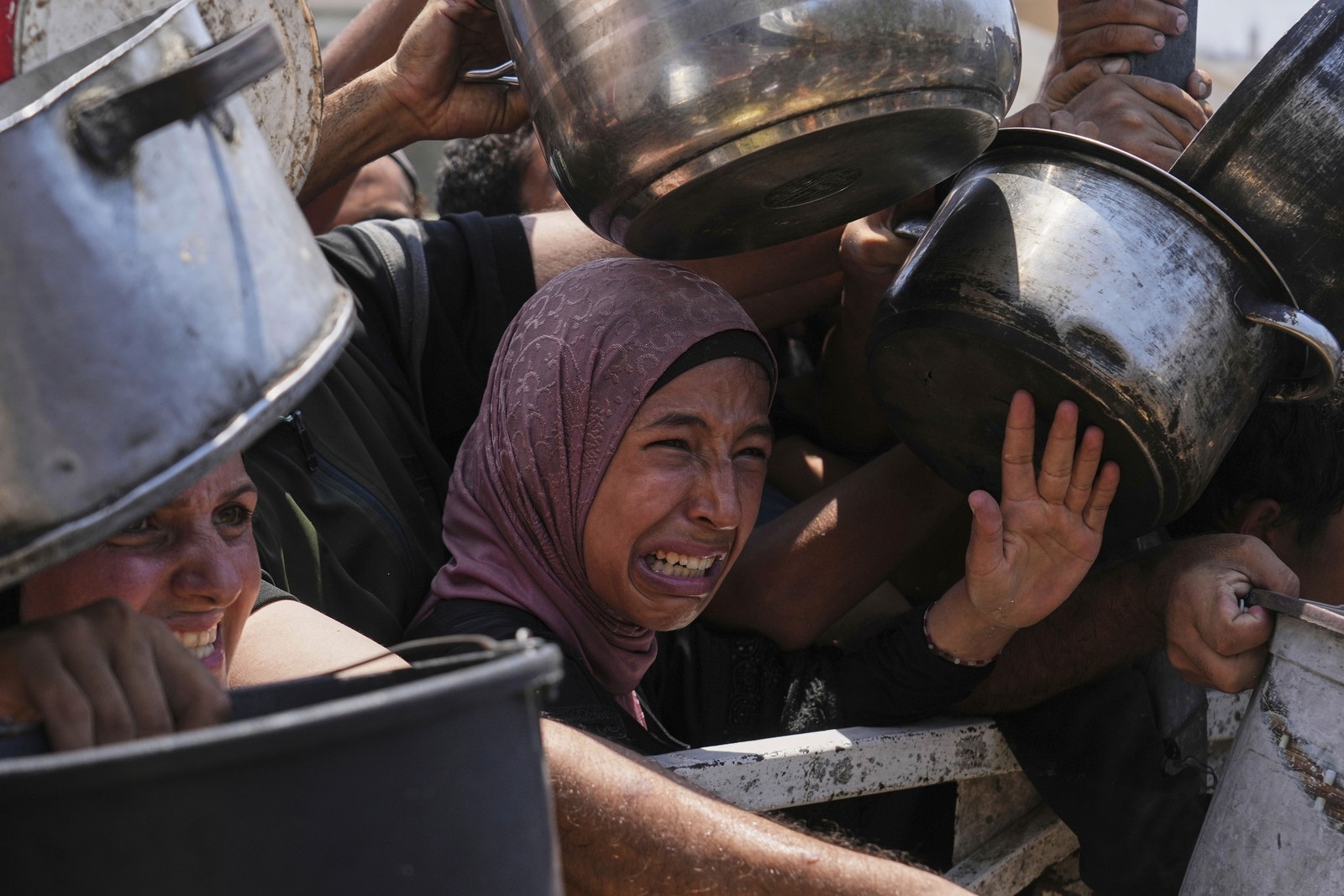 A close view of a person making an anguished face among a crowd holding out pots at a food distribution center