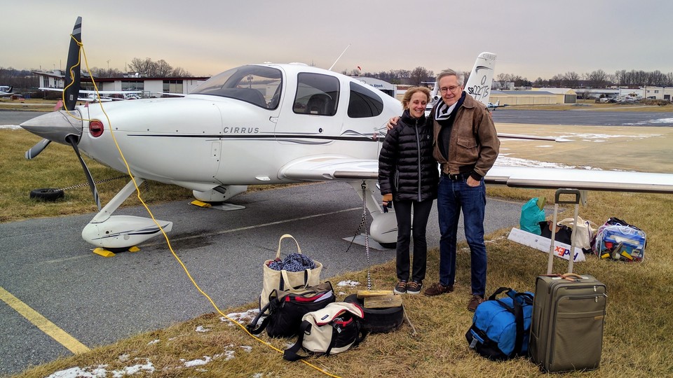 The authors, on a sub-freezing January 2017 morning at the Montgomery County Airpark, in Gaithersburg, Maryland, about to fly toward the west on the final leg of their previous trip. A new journey begins soon. (The yellow cord is to heat the engine sufficiently so it will start.) Around them is all the luggage their Cirrus SR22 would carry, for the next few months on the road.