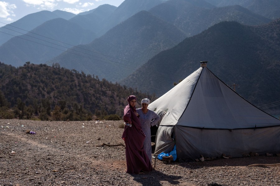 Two people stand outside a tent, with mountains rising in the background.