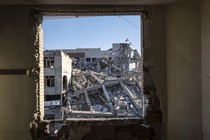 A photograph through a jagged hole in a wall shows a collapsed building across the street with an Iranian flag flying atop it.