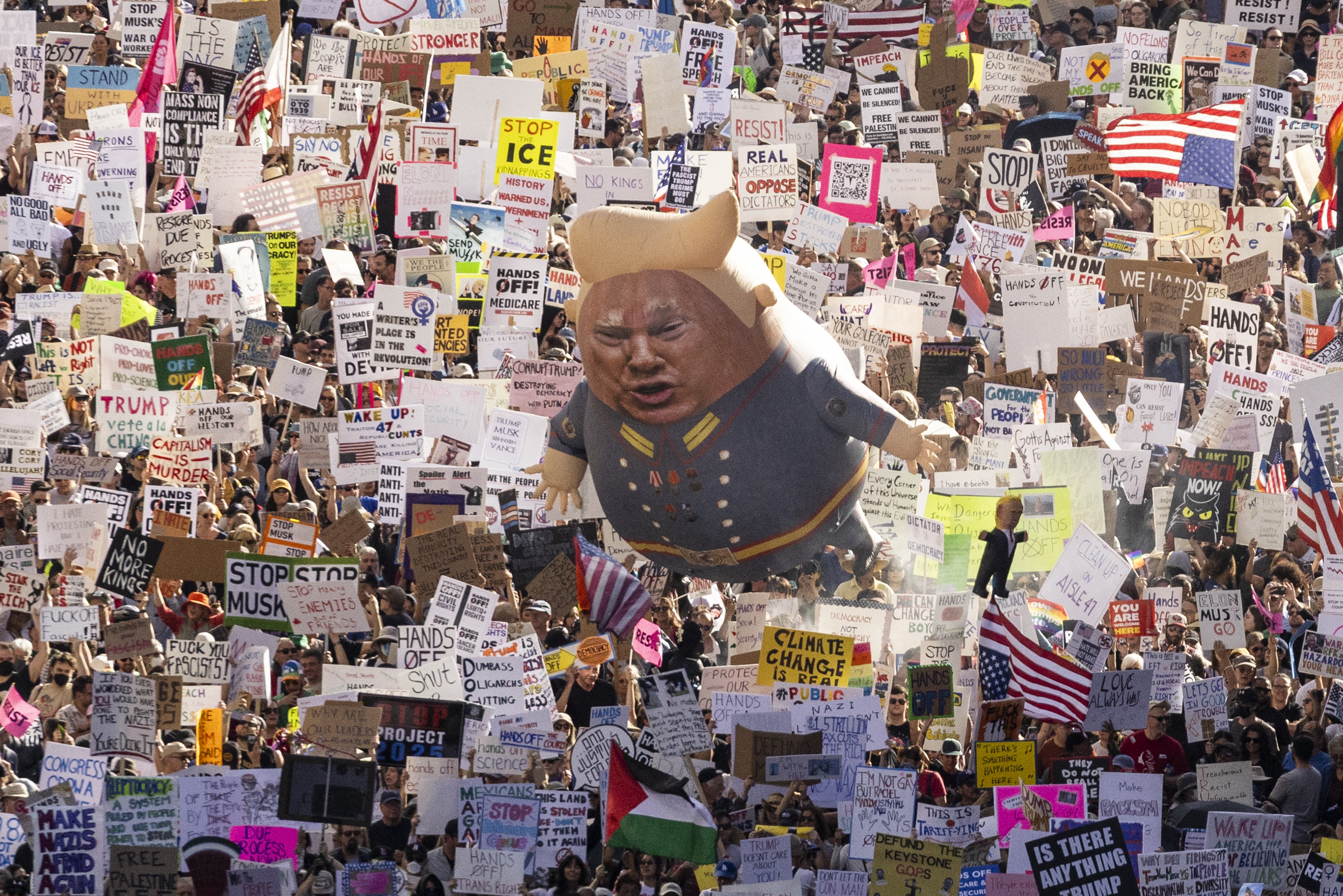 A large crowd of protesters marches down a Los Angeles street carrying many signs and a balloon with an image of President Donald Trump.