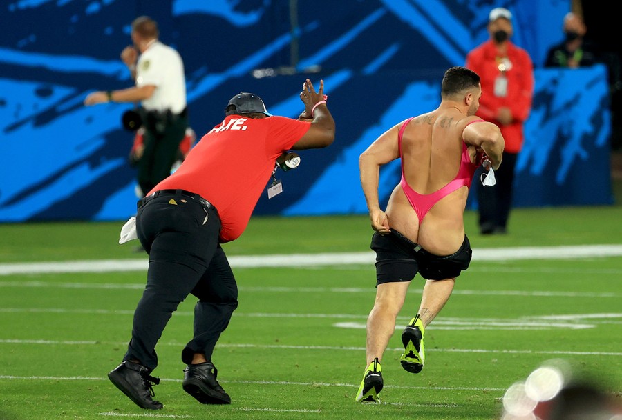 A man wearing very little pulls up his falling shorts as he runs from security on a football field.