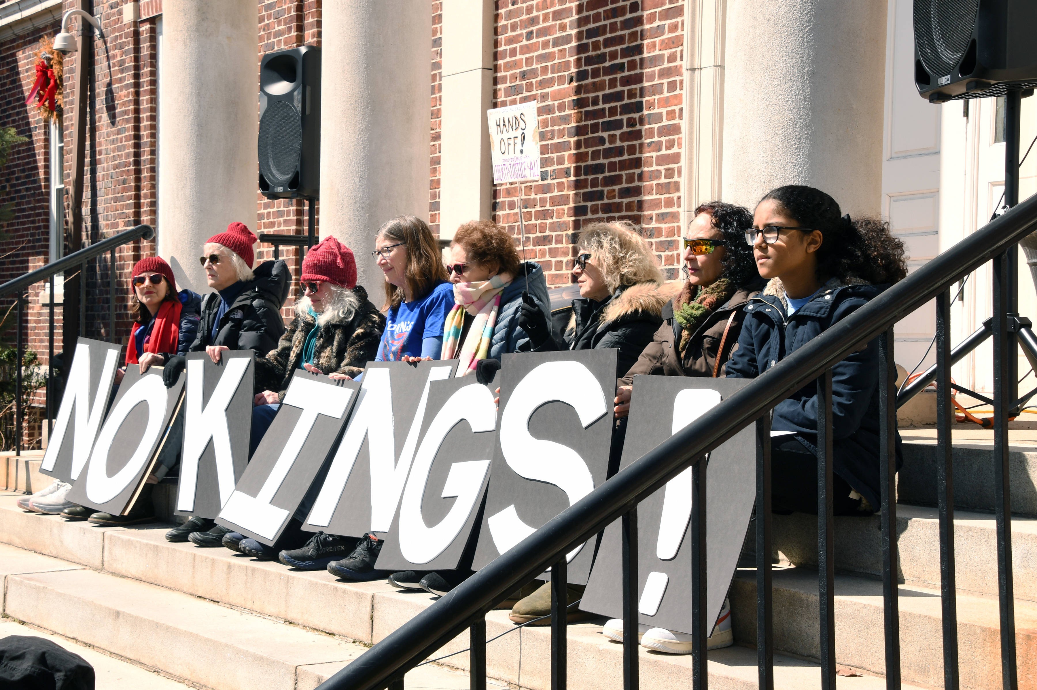 Eight women sit side-by-side on steps outside a building during a protest, holding up signs that, together, spell out 'no kings!'