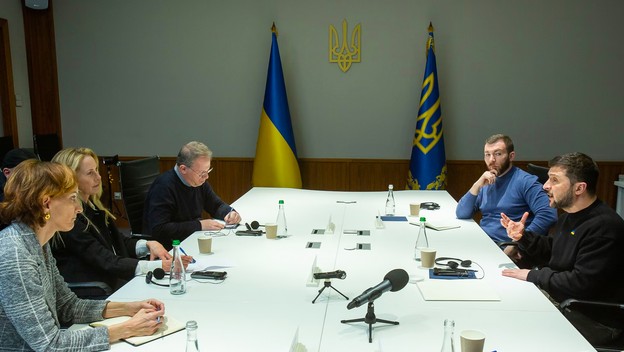 photo of large white meeting table with two women and a man on left and two men on right, one speaking, with Ukrainian flags in background