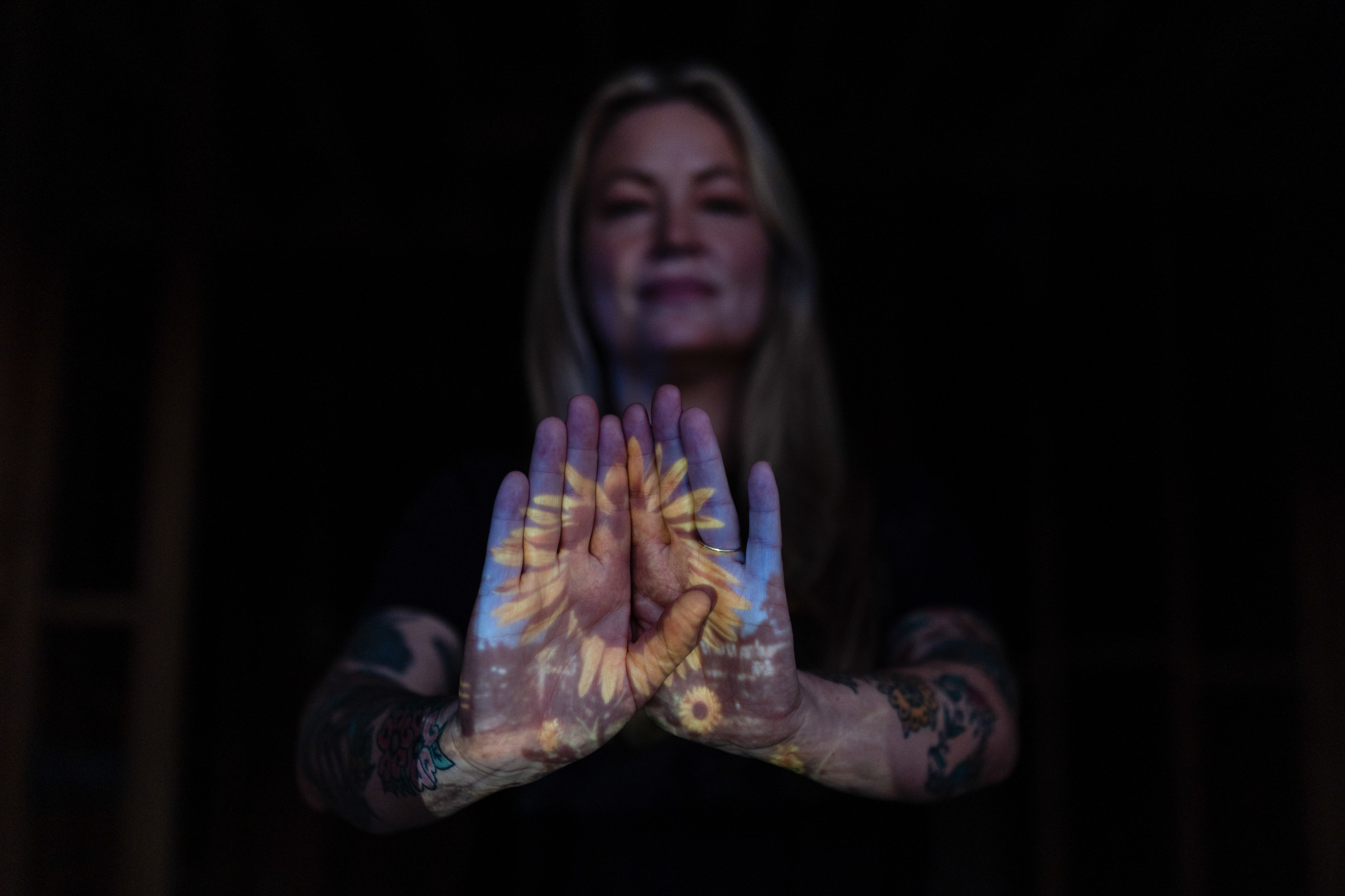 A woman poses, holding her hands up, with a projection of a photograph of a sunflower seen across her palms.