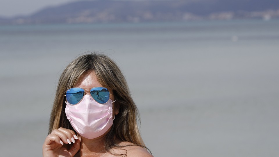 A woman on a beach in blue reflective sunglasses and a pink face mask