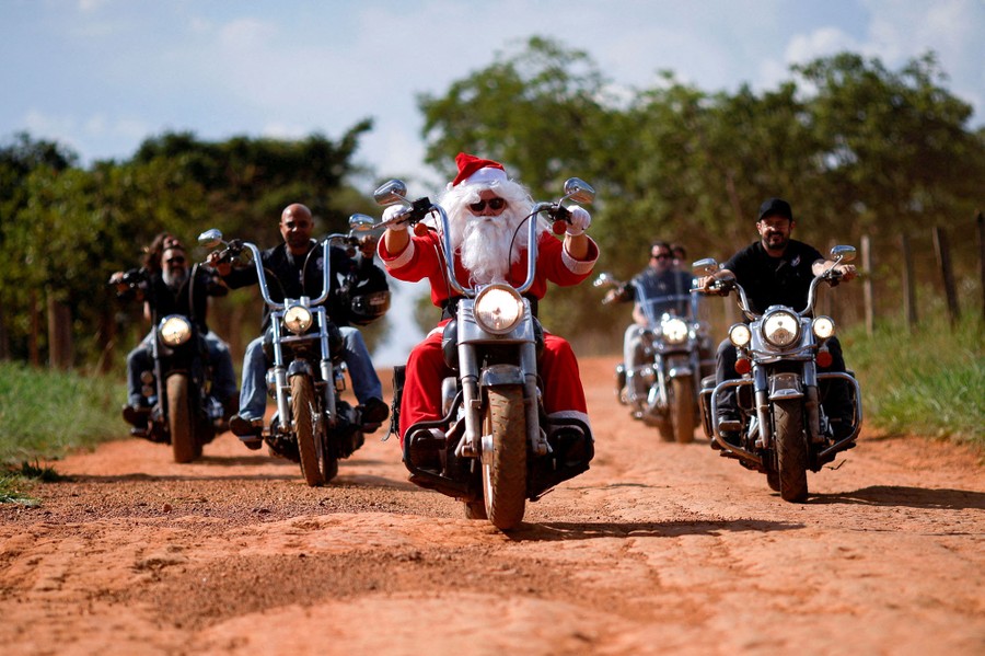 A man dressed as Santa Claus and several other men in regular clothes ride motorcycles on a dirt road.