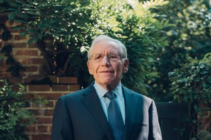 An older man in a blue suit and tie standing in front of a brick wall and lush greenery.
