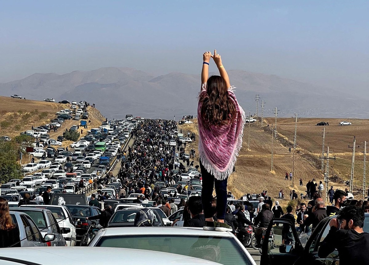 A woman holds her arms up, gesturing, while standing on a car among a large crowd of people marching past cars on a major road.