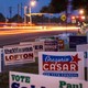 Political signs crowded together against the side of a road at night.