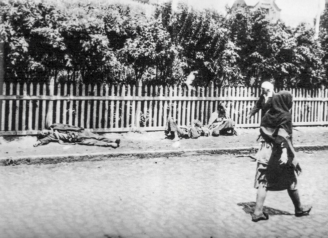 black and white photo of emaciated dying people lying on a sidewalk along a picket fence next to a street where people are walking
