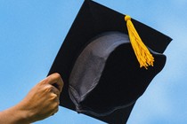 Photograph of a hand reaching out from the bottom left corner holding a mortarboard, as if it's about to be thrown