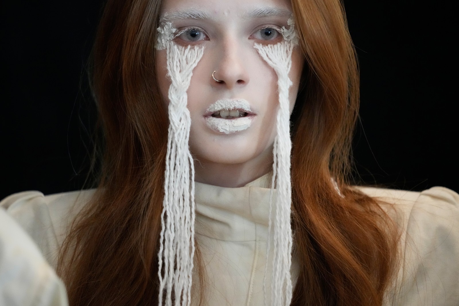 A model, seen during a fashion show, with long strings applied to her face, beneath each eye, creating an artistic image of flowing white tears.