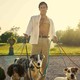 A man in an unbuttoned shirt stands holding the leashes of six dogs. He seems to be smirking at the camera.