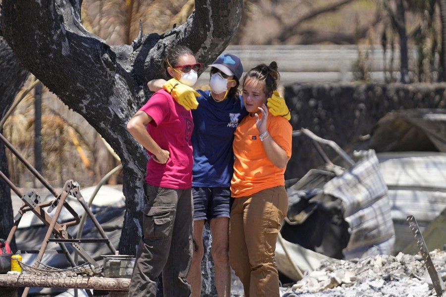 Three people embrace, standing amid the rubble of a burned house.