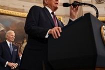 A photograph of Donald Trump speaking into a microphone, with Joe Biden in the background