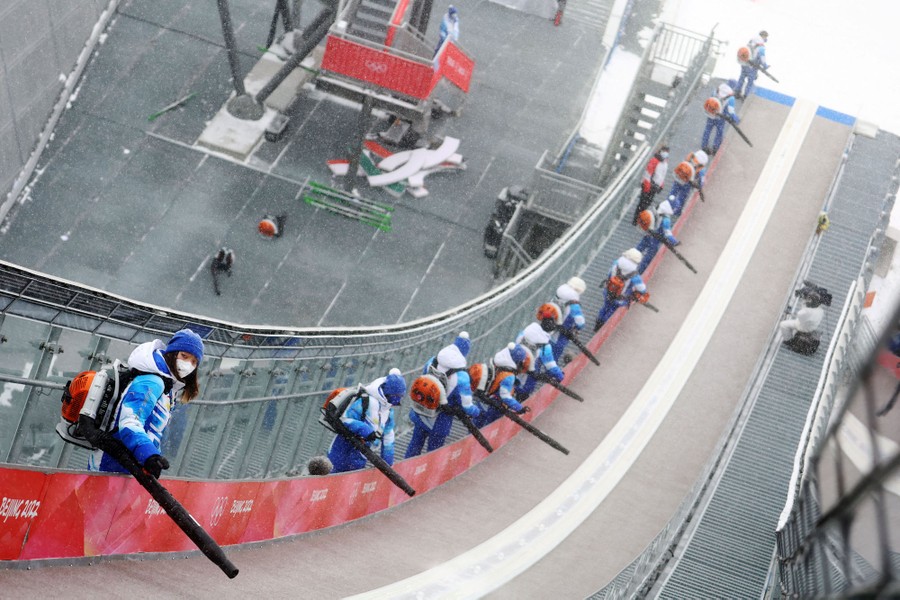 More than a dozen workers with leaf blowers clear snow from a ski ramp.