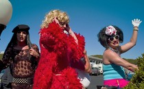 Three drag queens, one of whom is dressed in a long, red, feathered robe