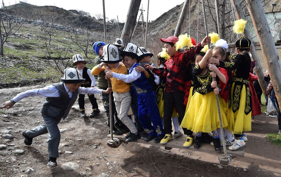 About a dozen young children wearing traditional clothing play on a large wooden swing.