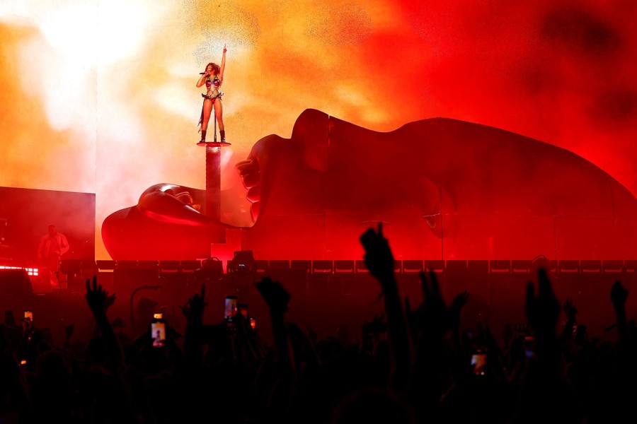 A performer sings on stage while standing on a platform above a giant sculpture of a human face with an open mouth.