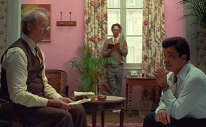 Three men, writers and editors of "The French Dispatch," in a pink room that features a large window, bold, floral-patterned drapes, and plants