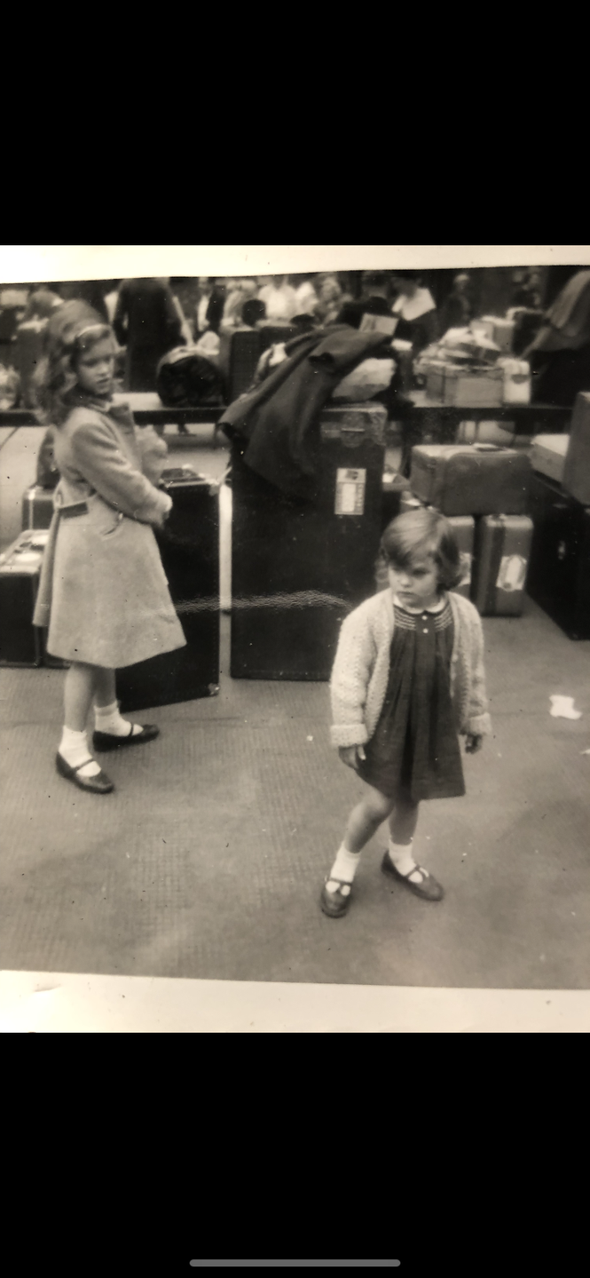 A black and white photo of a small girl (Caitlin Flanagan) wearing a dress, cardigan, and Mary Jane shoes, standing in front of suitcases, while another young girl wearing a coat and Mary Jane shoes looks on 