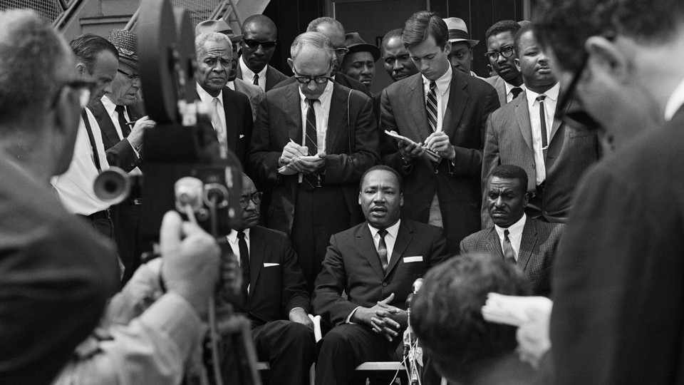 Martin Luther King Jr. speaks in front of news cameras and reporters with notepads, flanked by Fred Shuttlesworth and L.H. Pitts.