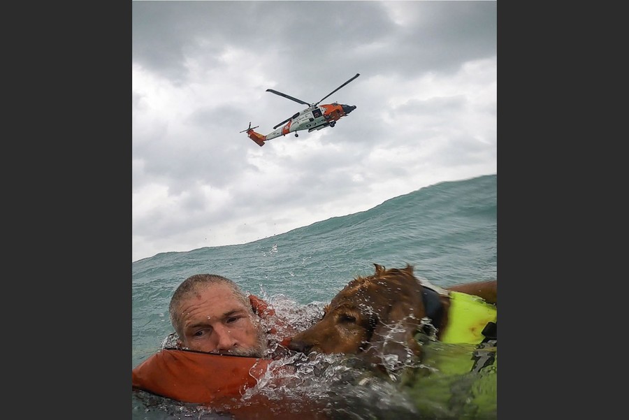 A man and his dog float in rough ocean water, snapping a selfie as a rescue helicopter flies overhead.