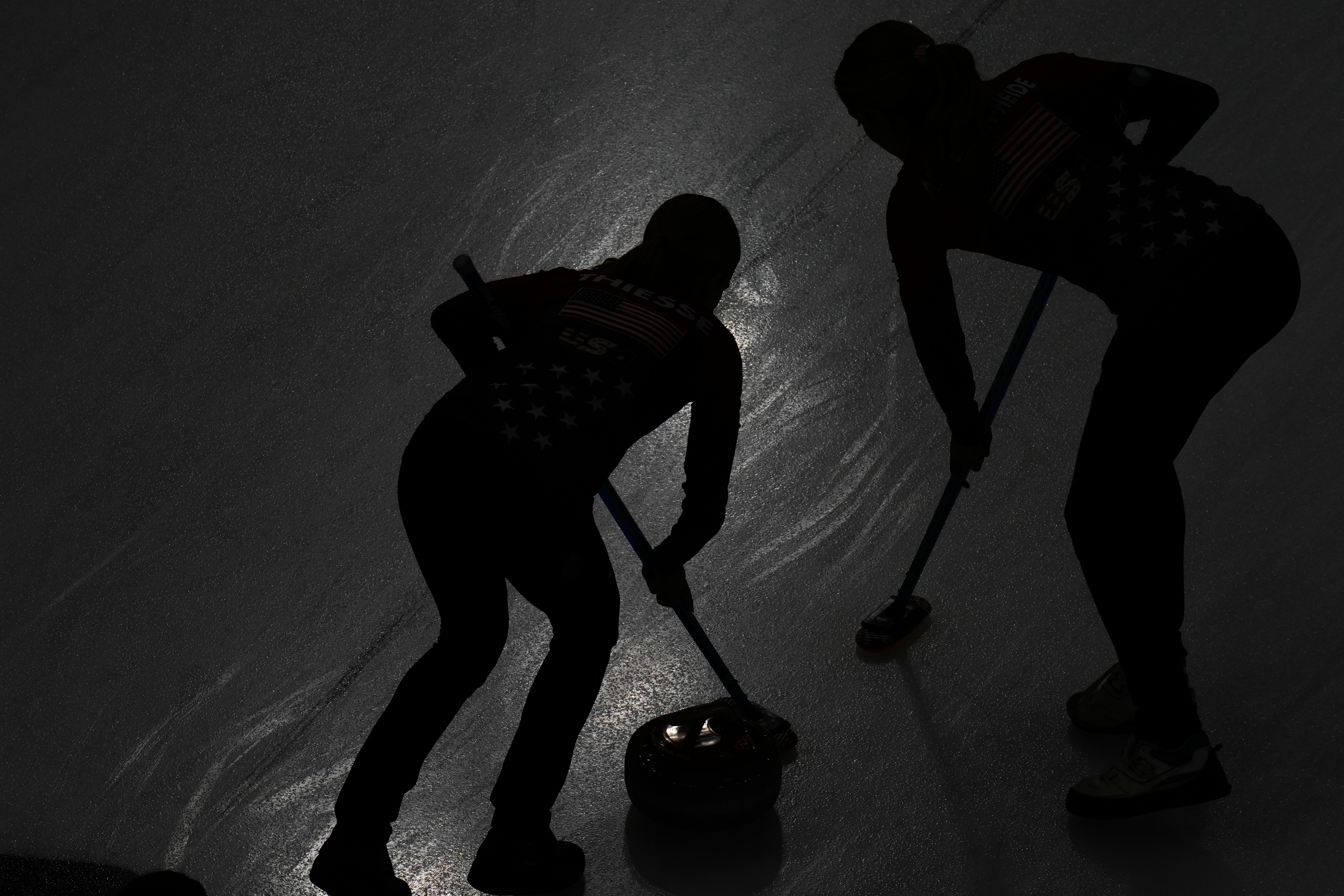 Two athletes compete during a curling match, seen in silhouette on ice.