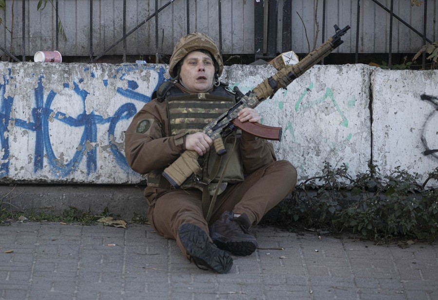 A soldier sits on a curb, leaning on a barrier, holding his weapon.