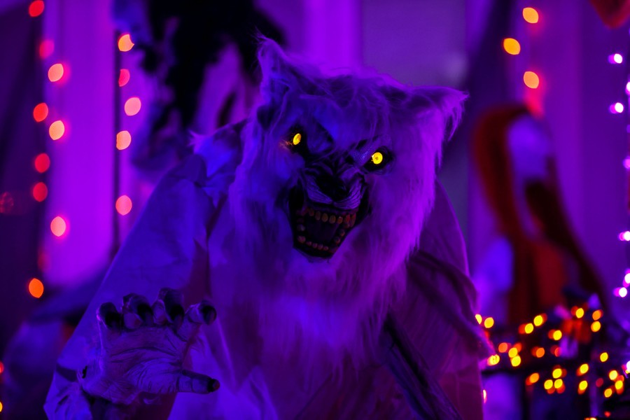 A large figure of a wolf-man with illuminated eyes stands in front of a decorated house.