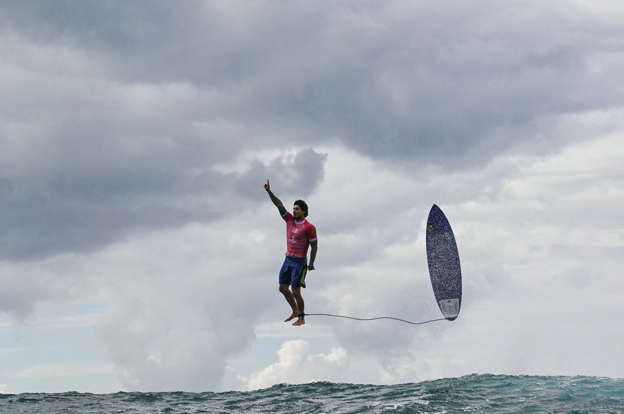 A surfer points upward while jumping above a wave with his surfboard beside him, appearing to stand casually upright in midair.