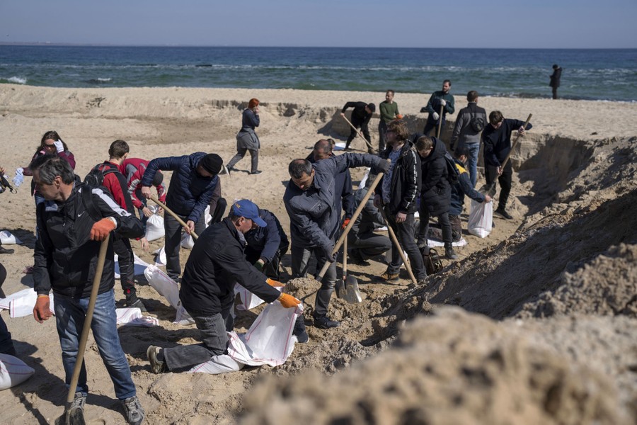 About two dozen people are working together with shovels and sacks on a beach, making sandbags.