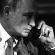 A black-and-white photo of Vladimir Putin holding a phone to his left ear