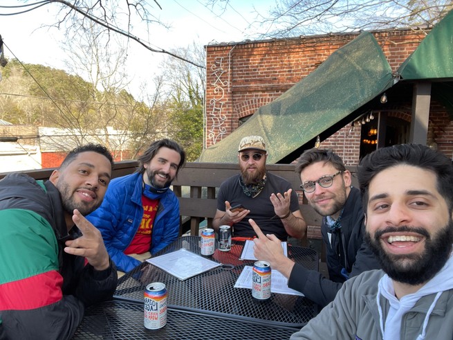 Five men sit outside around a patio table, dressed in warm coats, smiling for the camera