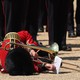 A soldier in dress uniform, holding a trombone, lies on the ground after falling.