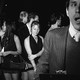 Black-and-white photo of people lining up in front of a nightclub next to a man in a striped suit holding a clipboard