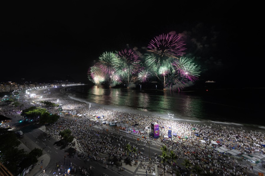 A large crowd watches fireworks from a broad, curving beach.