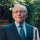 An older man in a blue suit and tie stands in front of a brick wall and lush greenery