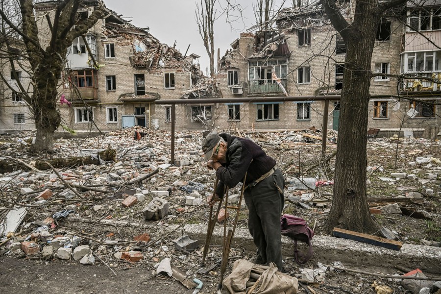 A man leans on crutches in a street in front of an apartment building that has been blown apart.