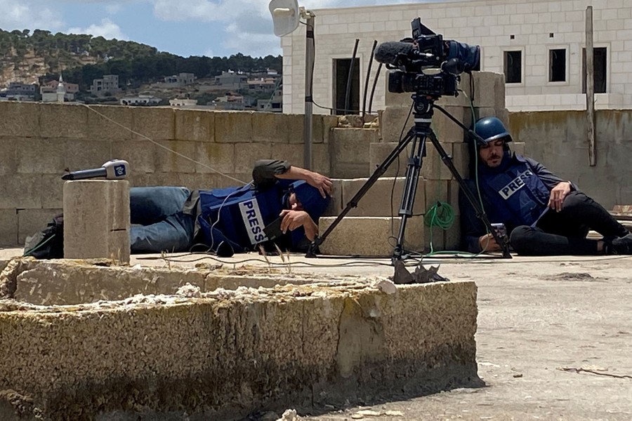 Two journalists wearing helmets and bulletproof vests that say "Press" crouch low, behind a wall, on a rooftop.