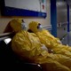 Two hospital workers in yellow hazmat suits rest on chairs in a dimly lit hallway