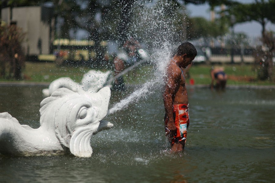 A person stands in a fountain, being splashed by water coming from a fish sculpture.
