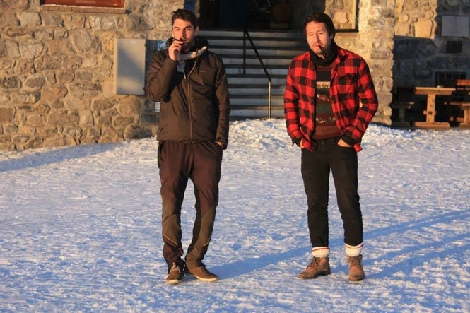 Two men stand in the snow outside a stone building, with cigars in their mouth.