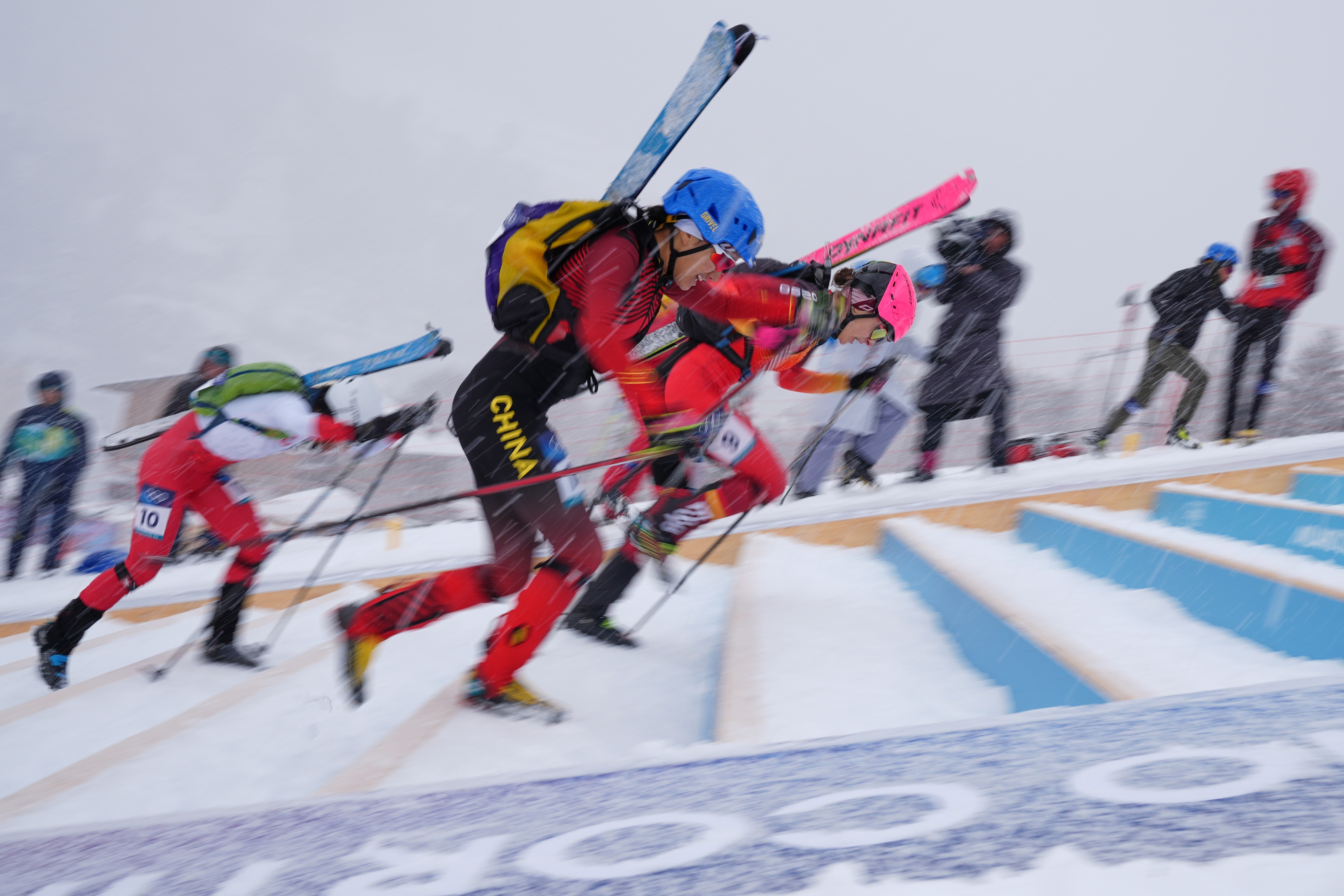 Three racers carrying skis on their backs run up a set of steps laid into a snow-covered mountain slope.
