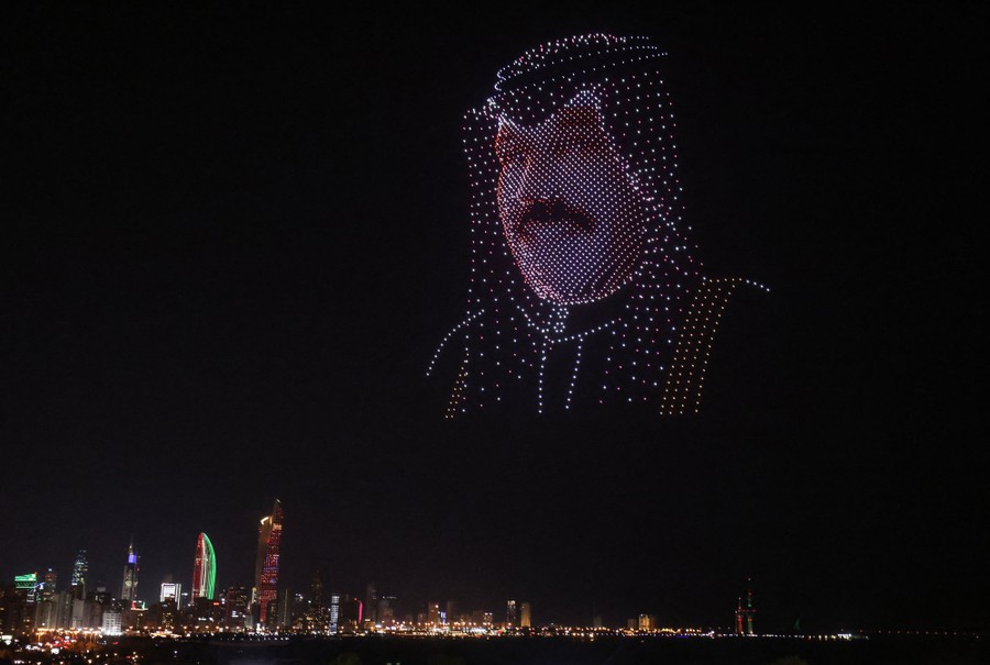 Dozens of illuminated drones hover in a coordinated manner to form the image of a man's head and face in the night sky.