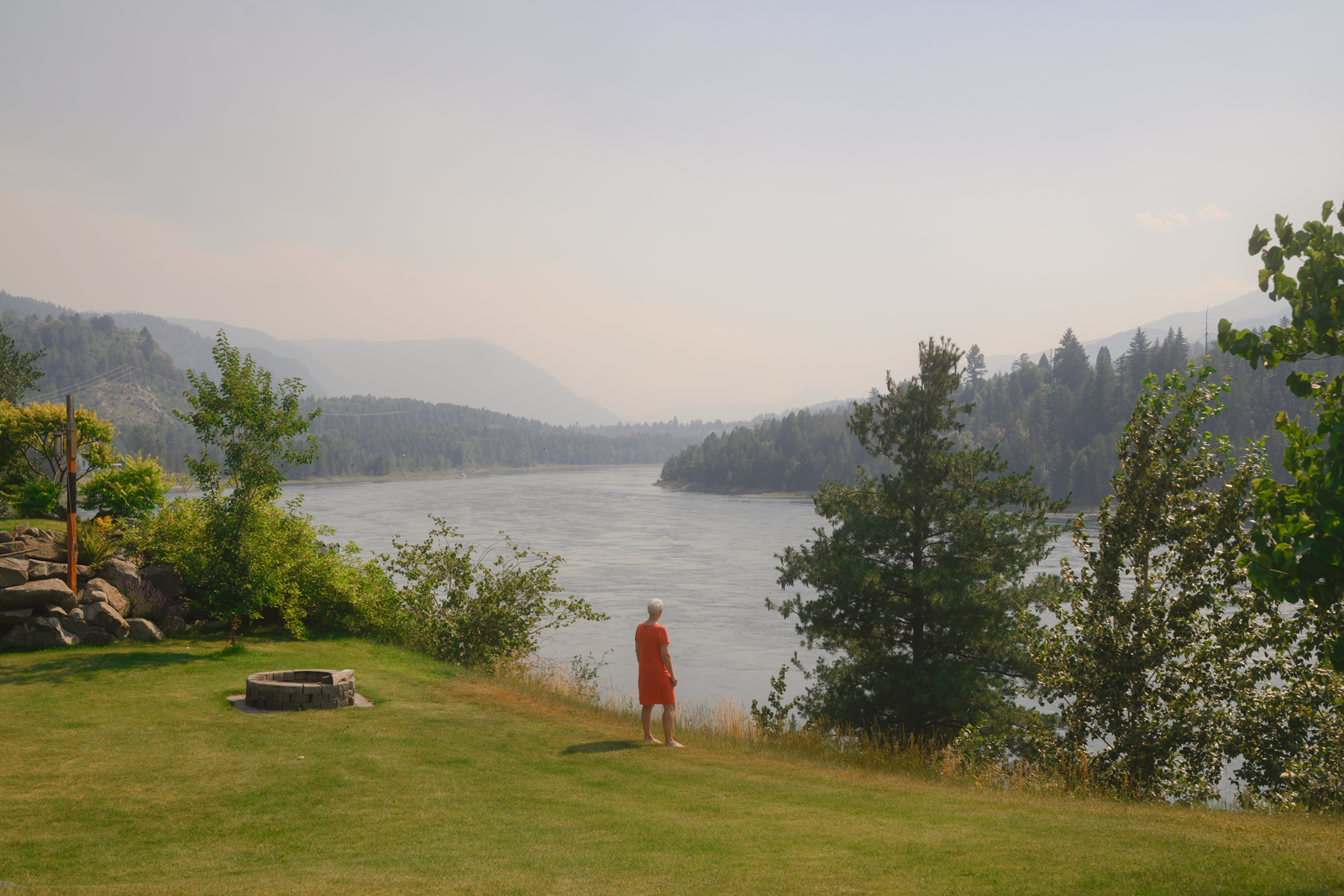 photo of person in red standing on green lawn and looking at river with distant mountains and forest in background