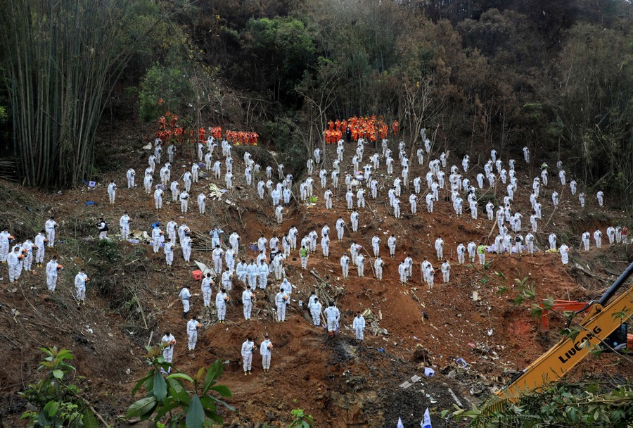 Dozens of rescue workers in white protective gear stand on a disturbed hillside, the site of a plane crash.