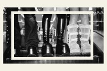 A black-and-white image of three pairs of boots being worn by people, standing on an escalator step