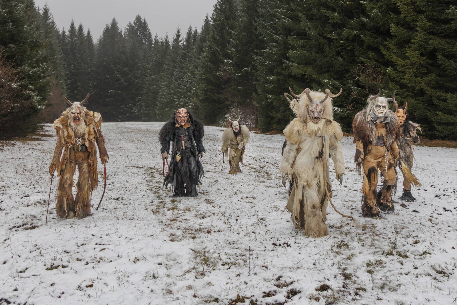 Six performers wearing frightening Krampus costumes and masks walk across a snow-covered field out of a forest.